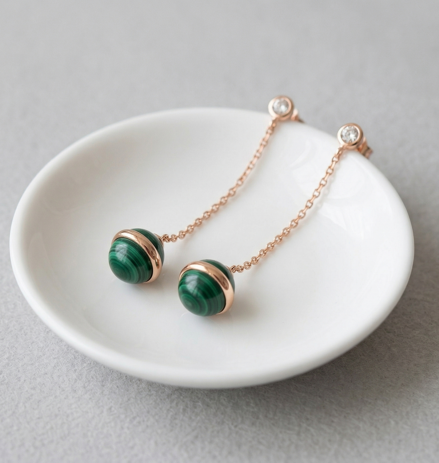 Pair of natural malachite drop earrings resting in white ceramic jewelry dish, rose gold chain and CZ stud visible, light gray background