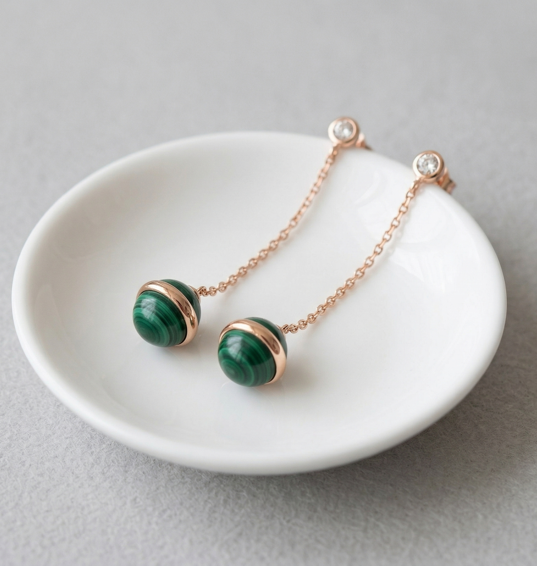 Pair of natural malachite drop earrings resting in white ceramic jewelry dish, rose gold chain and CZ stud visible, light gray background