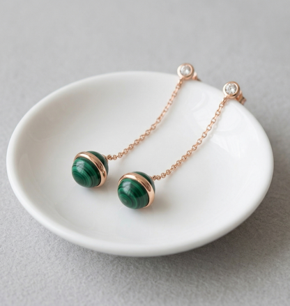 Pair of natural malachite drop earrings resting in white ceramic jewelry dish, rose gold chain and CZ stud visible, light gray background