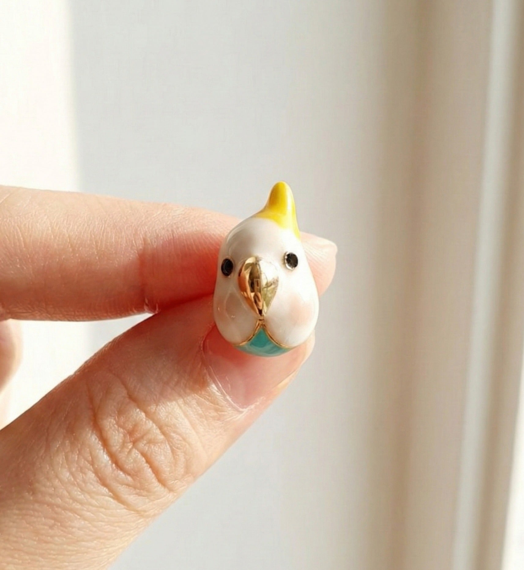  Single cockatiel earring held between fingers — yellow crest and gold beak detail visible 