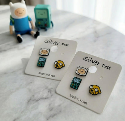 Flat lay Adventure Time earrings Finn Jake BMO on white packaging with character toys in background