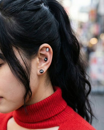 Model wearing No-Face and soot sprite stud earrings styled with red turtleneck sweater, Ghibli-themed ear stack with multiple character piercings on helix, conch, and lobe, full ear view showcasing versatile piercing placement.