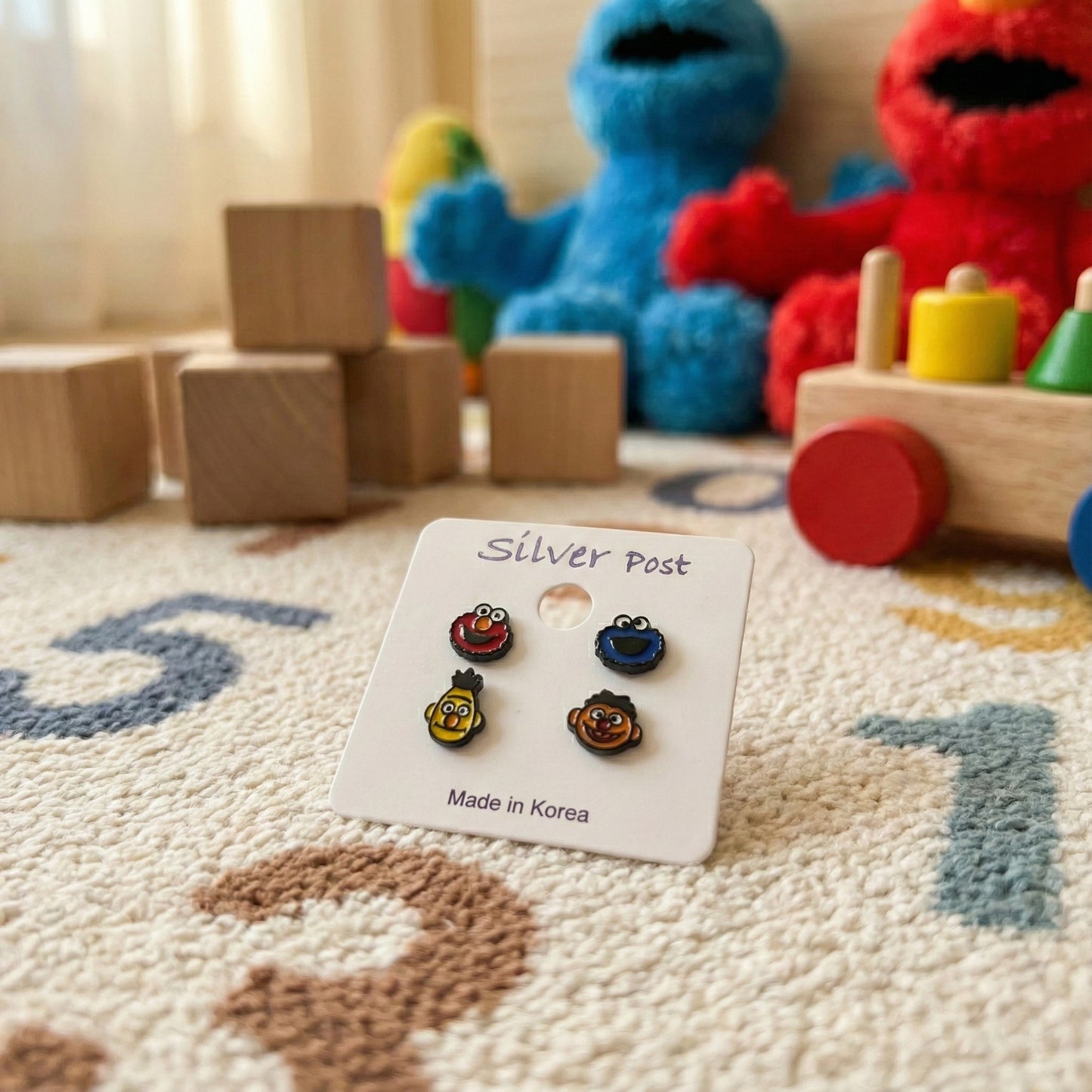 Sesame Street character earrings set on white card with plush toys in background showcasing product packaging
