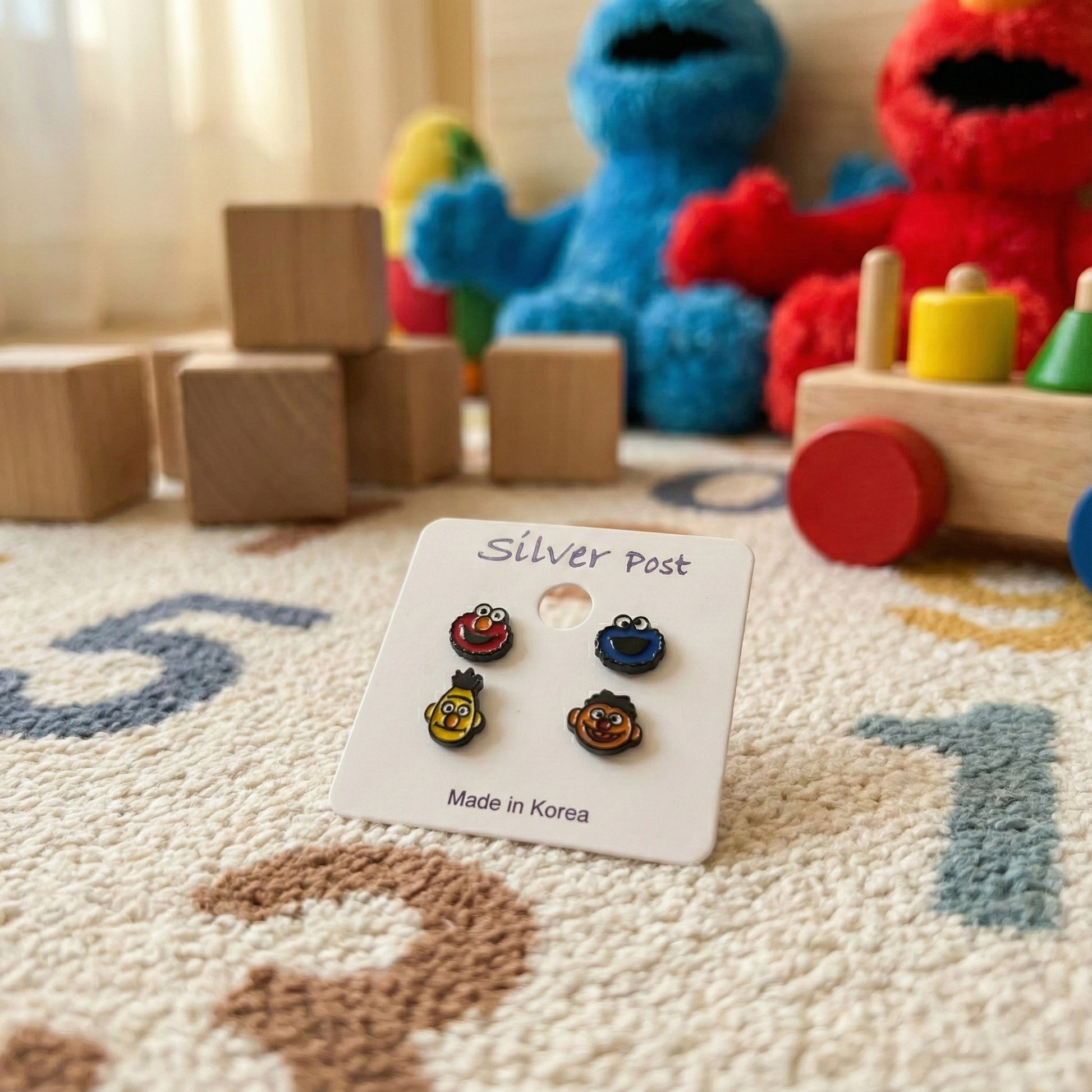 Sesame Street character earrings set on white card with plush toys in background showcasing product packaging