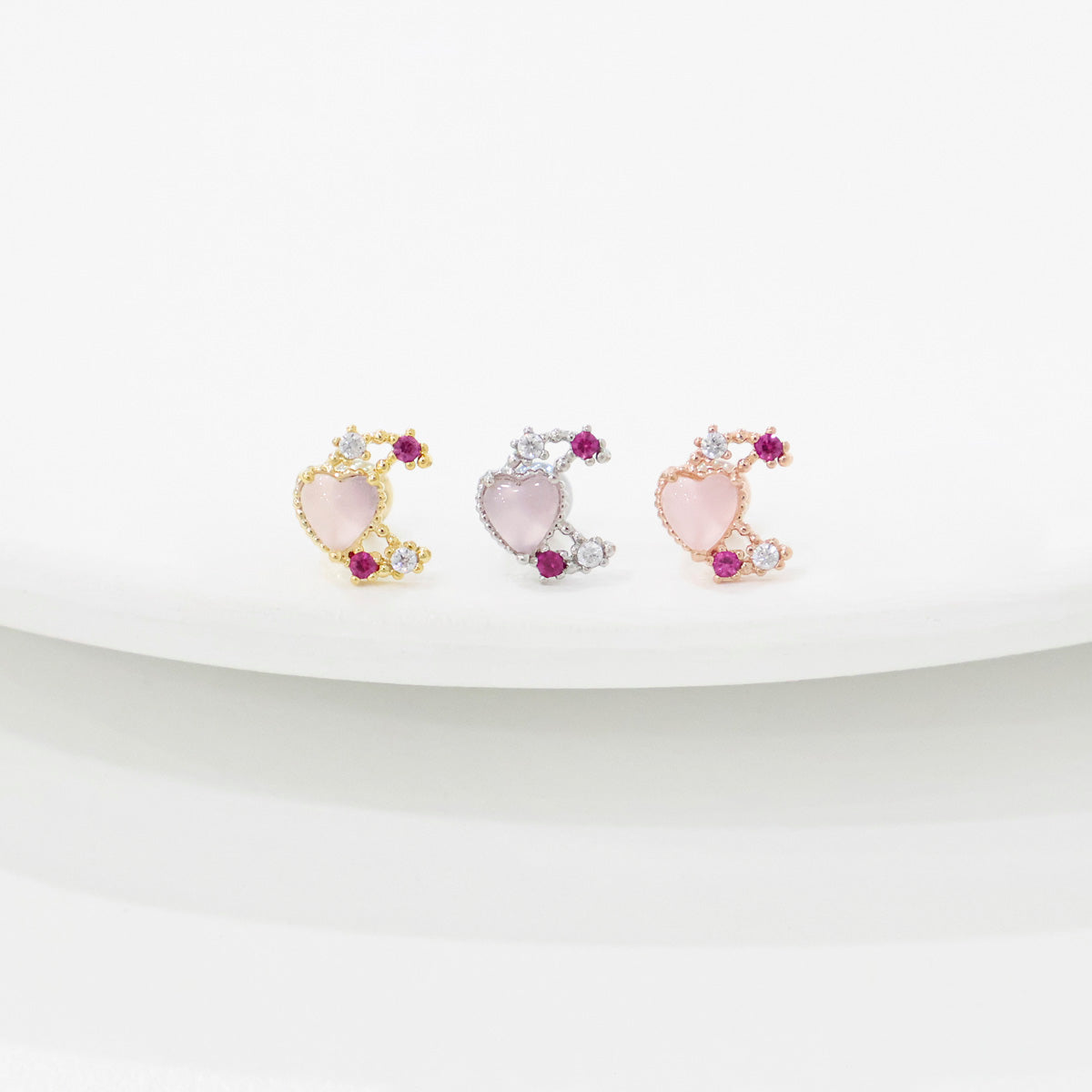 Three pink rose quartz moon heart barbell studs in gold, silver, and rose gold plating on white curved surface