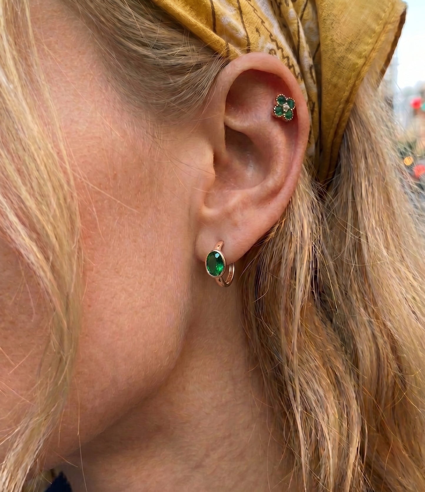 Detailed close-up of a model's ear featuring a vibrant emerald green oval gemstone earring in a secure bezel setting, paired with a matching green crystal clover cartilage piercing. She is wearing a vintage yellow paisley pattern bandana headband, creating a retro and stylish look.