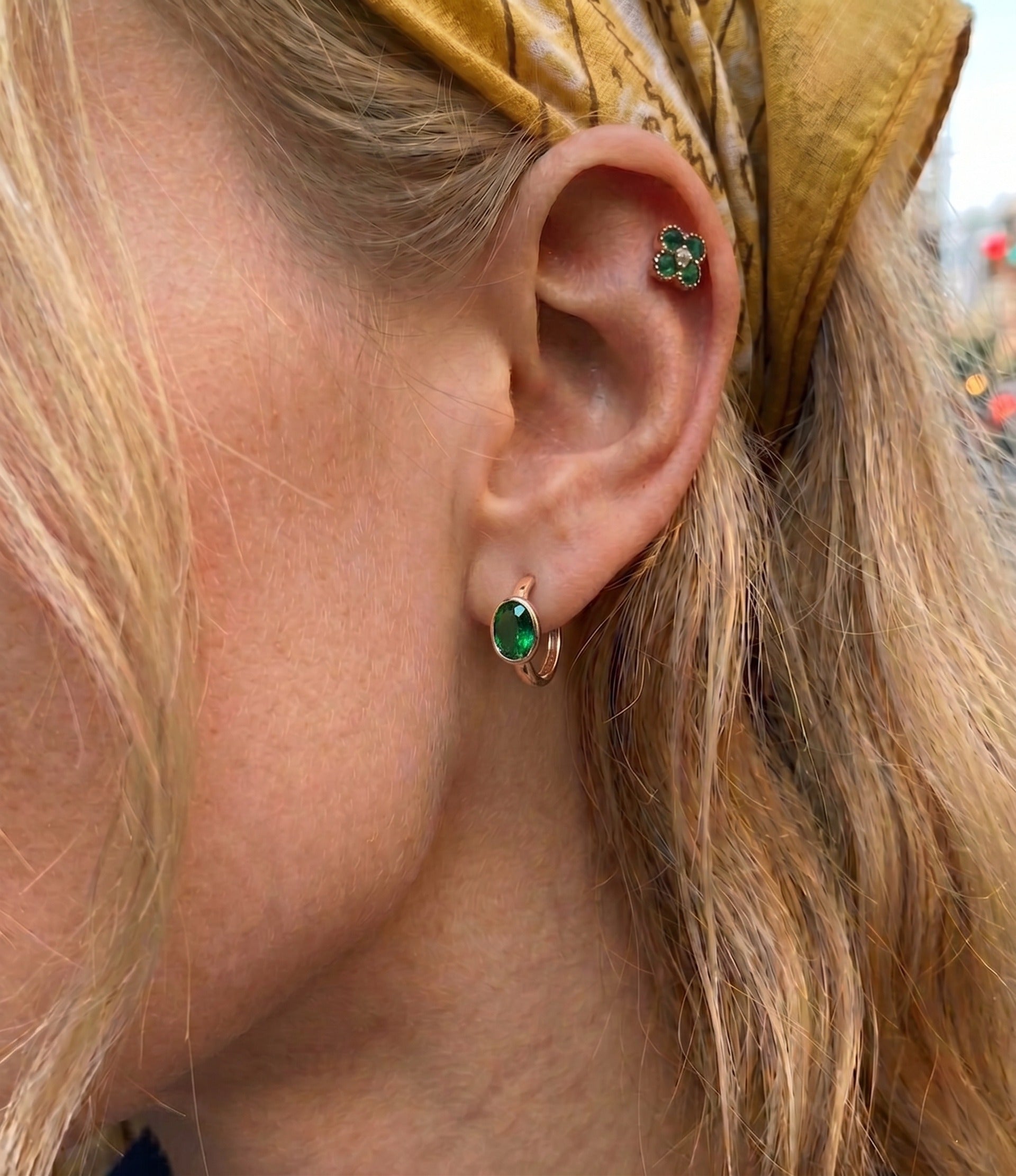 Detailed close-up of a model's ear featuring a vibrant emerald green oval gemstone earring in a secure bezel setting, paired with a matching green crystal clover cartilage piercing. She is wearing a vintage yellow paisley pattern bandana headband, creating a retro and stylish look.