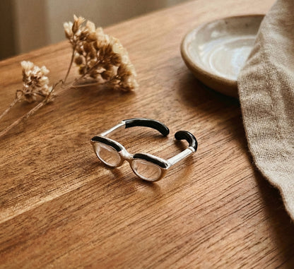 Flat lay of silver spectacle ring showing side view with black enamel temple tips on white stone surface