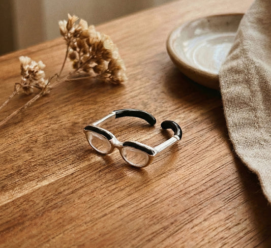 Flat lay of silver spectacle ring showing side view with black enamel temple tips on white stone surface