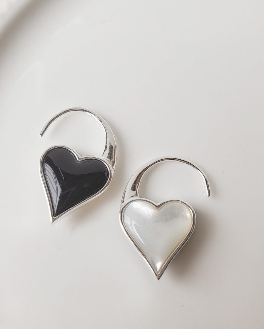 Flat-lay of silver hook earrings with heart pendants in glossy black onyx and luminous mother-of-pearl, on clean white background.