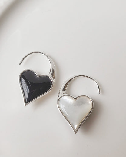 Flat-lay of silver hook earrings with heart pendants in glossy black onyx and luminous mother-of-pearl, on clean white background.