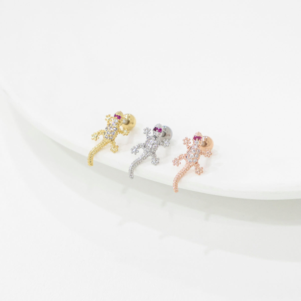 Three lizard-shaped earrings in gold, silver, and rose gold on a white surface.