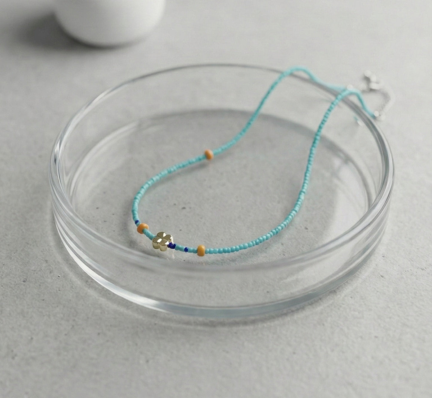 Turquoise blue seed bead necklace with gold four-leaf clover charm and orange accent beads on glass dish