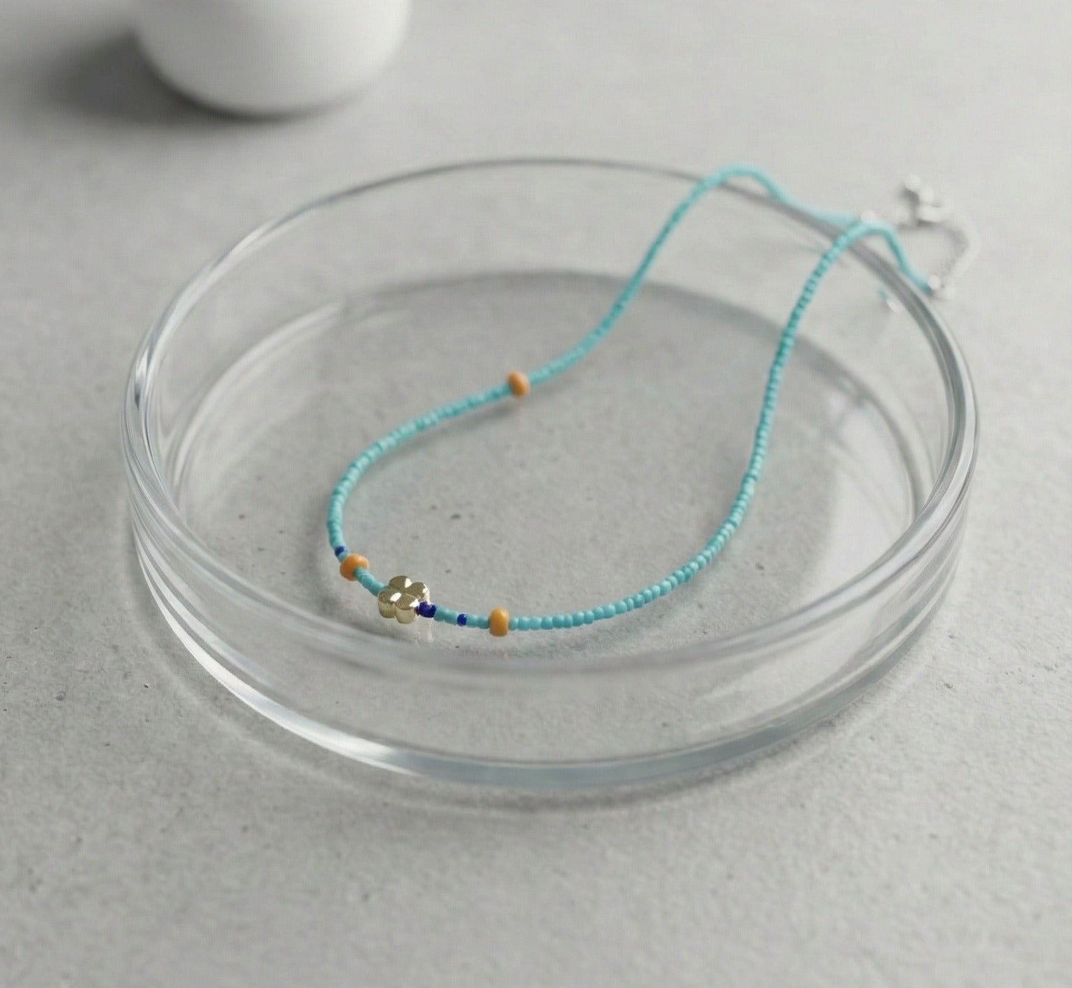 Turquoise blue seed bead necklace with gold four-leaf clover charm and orange accent beads on glass dish