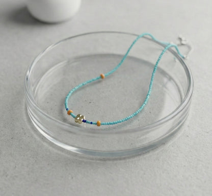 Turquoise blue seed bead necklace with gold four-leaf clover charm and orange accent beads on glass dish