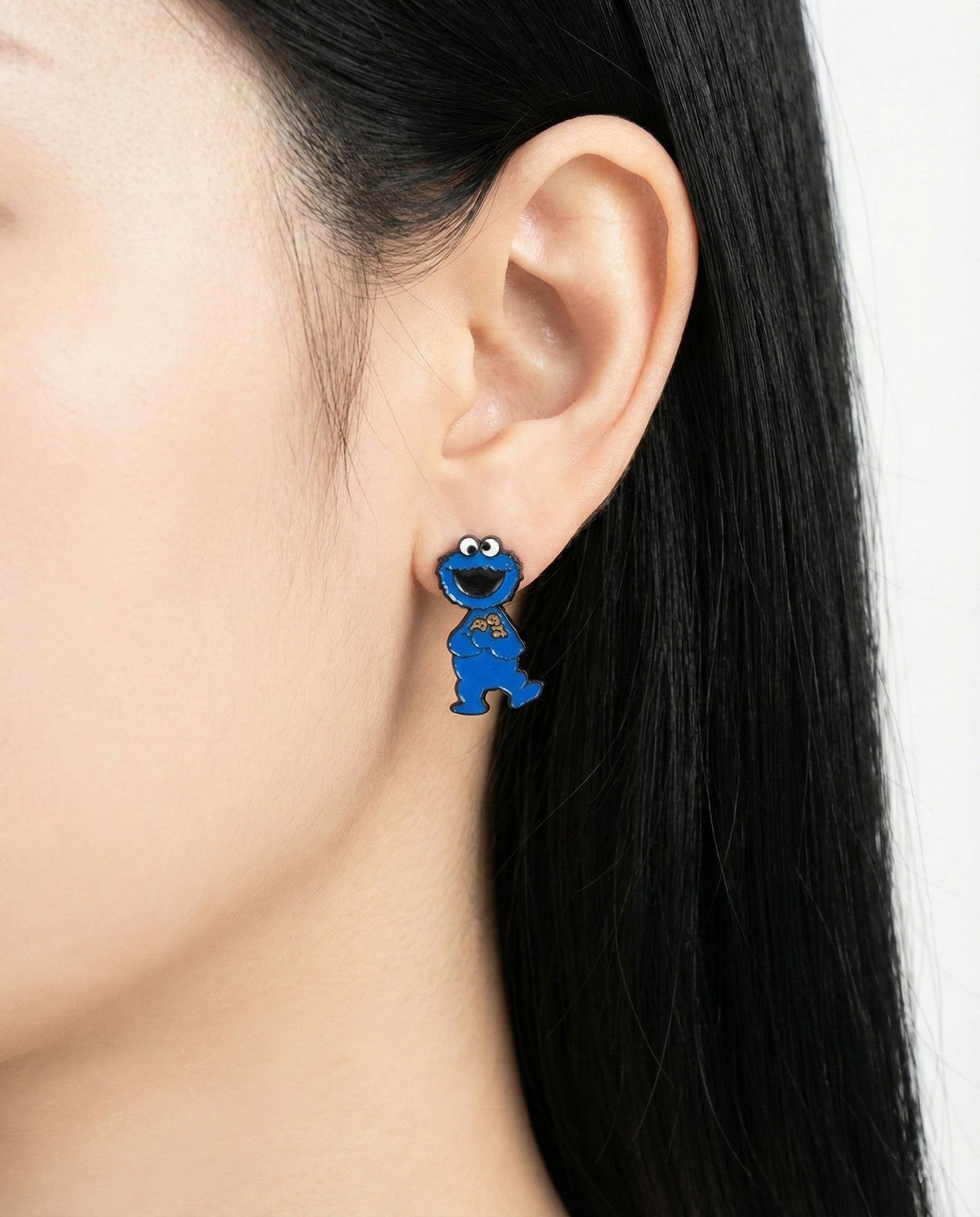 Cookie Monster full-body silver post earring worn on earlobe, vibrant blue design standing out on dark hair.