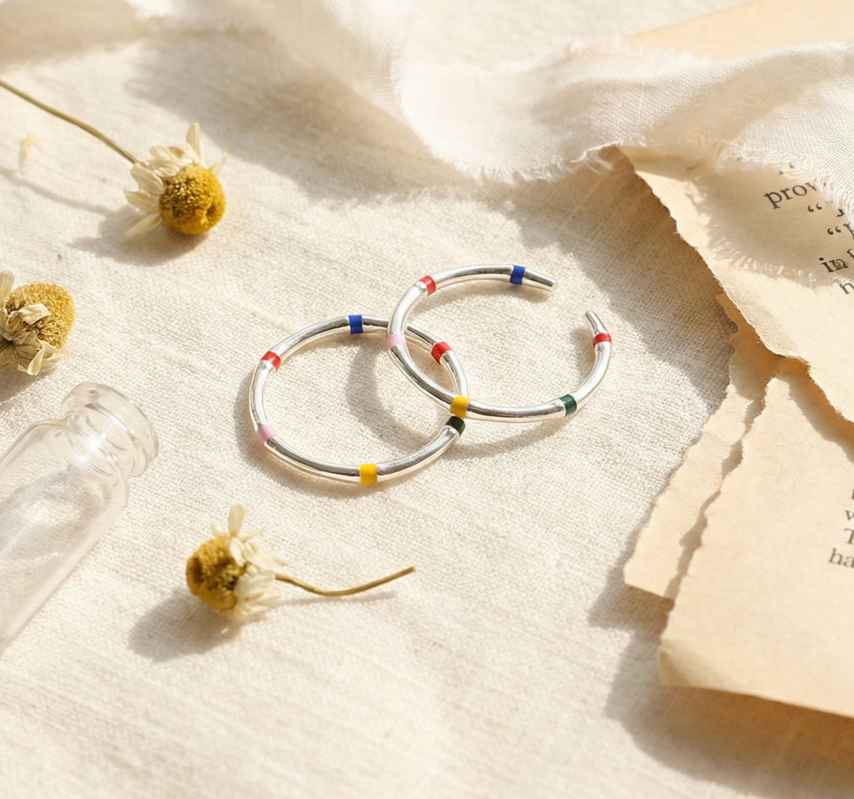 Flat lay of two 925 sterling silver multicolor enamel open rings with dried flowers and vintage book pages, lifestyle product photography