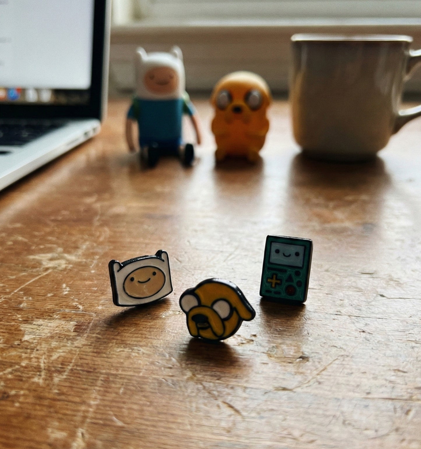 Desk styling with Adventure Time stud earrings Finn Jake BMO displayed on wooden surface with laptop