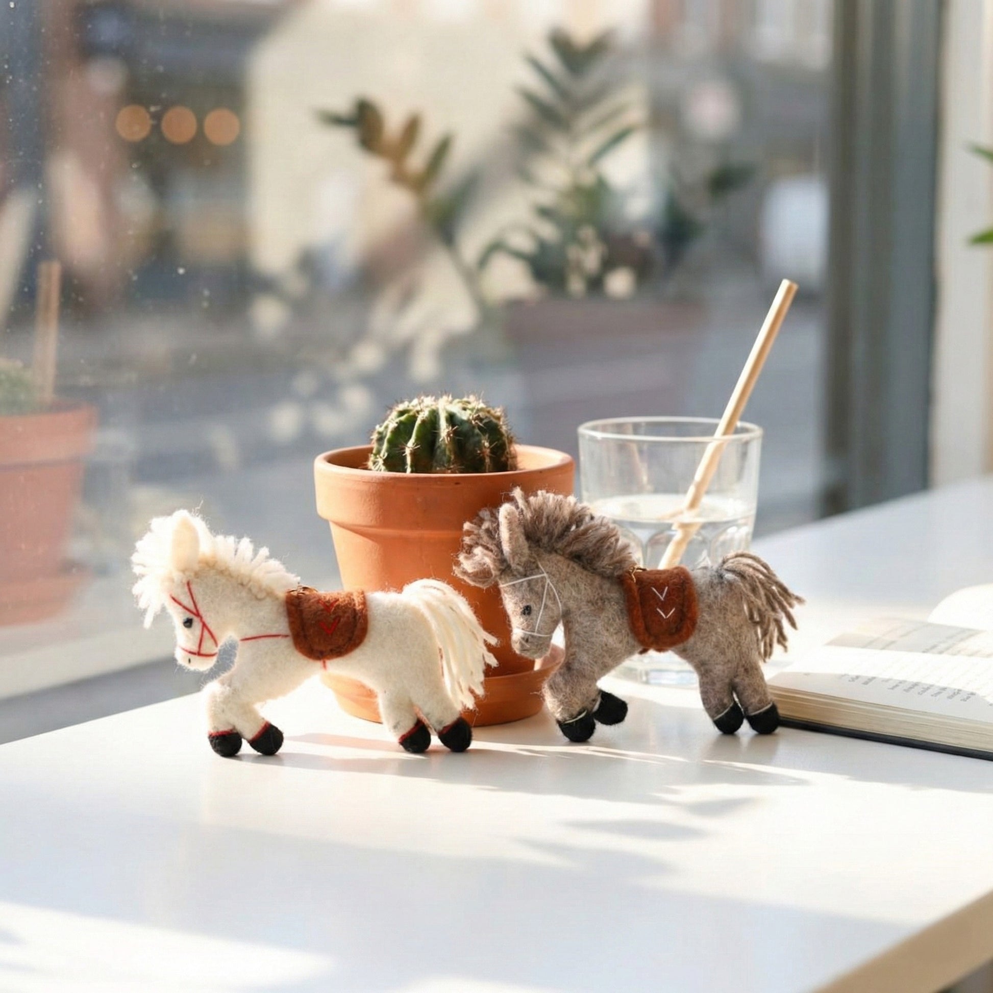 White and grey wool felt horse charms displayed on cafe table with cactus plant and glass