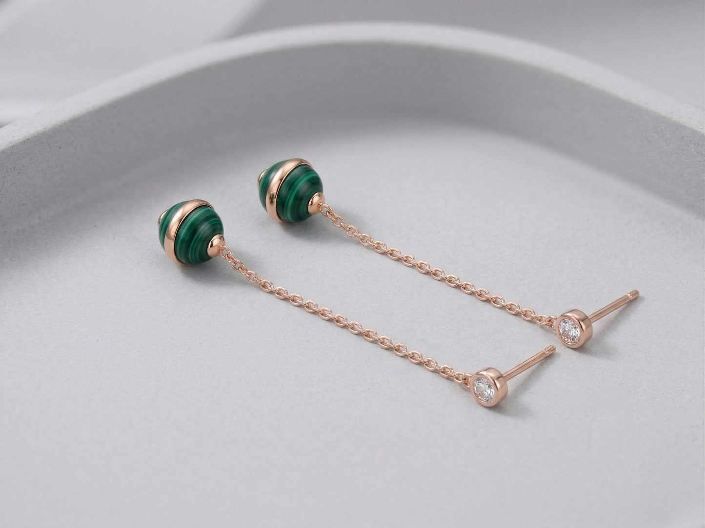 Two malachite ball drop earrings laid flat on pale gray curved surface, showing rose gold push-back stud posts and full chain length detail