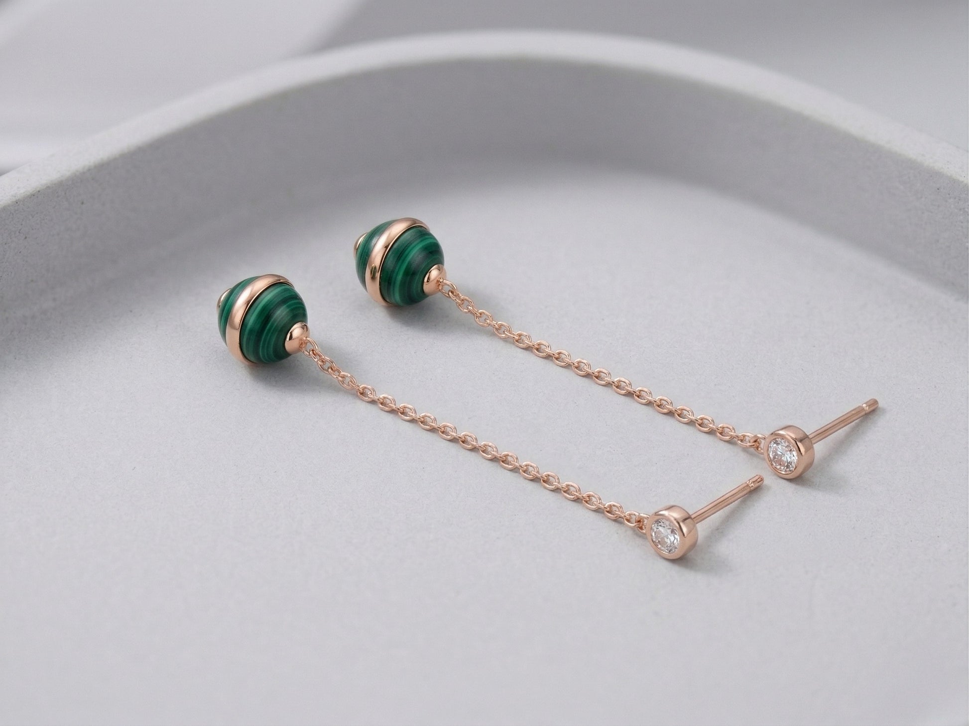 Two malachite ball drop earrings laid flat on pale gray curved surface, showing rose gold push-back stud posts and full chain length detail