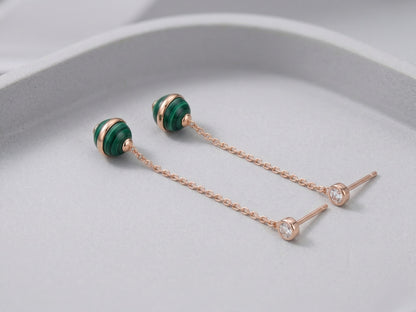 Two malachite ball drop earrings laid flat on pale gray curved surface, showing rose gold push-back stud posts and full chain length detail