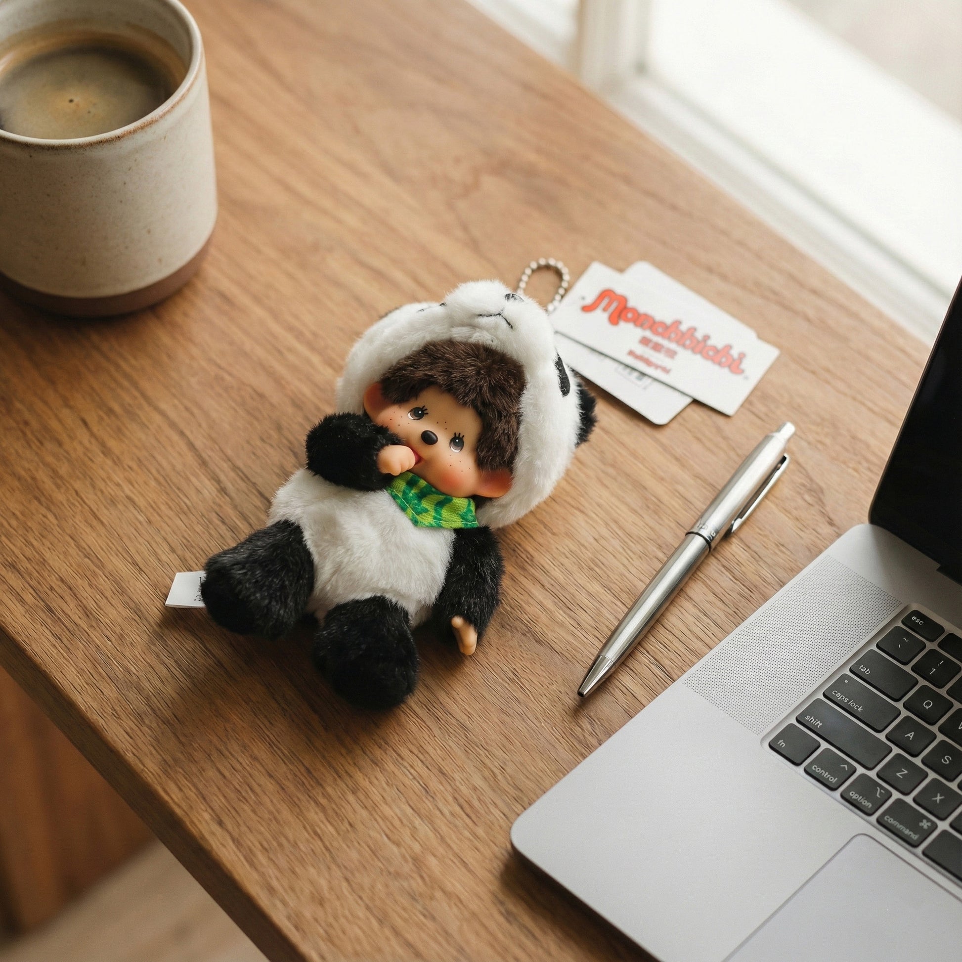 Panda Monchhichi plush keychain on wooden desk with laptop and coffee