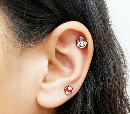 Mario face stud worn on the upper ear and a red mushroom stud on the lobe.