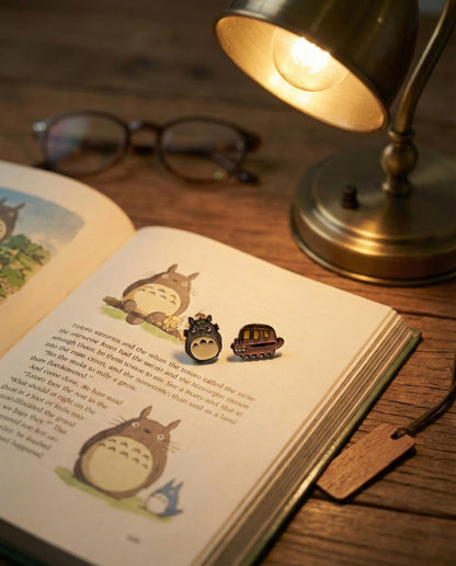 Totoro and Catbus stud earrings on open Studio Ghibli storybook with warm desk lamp