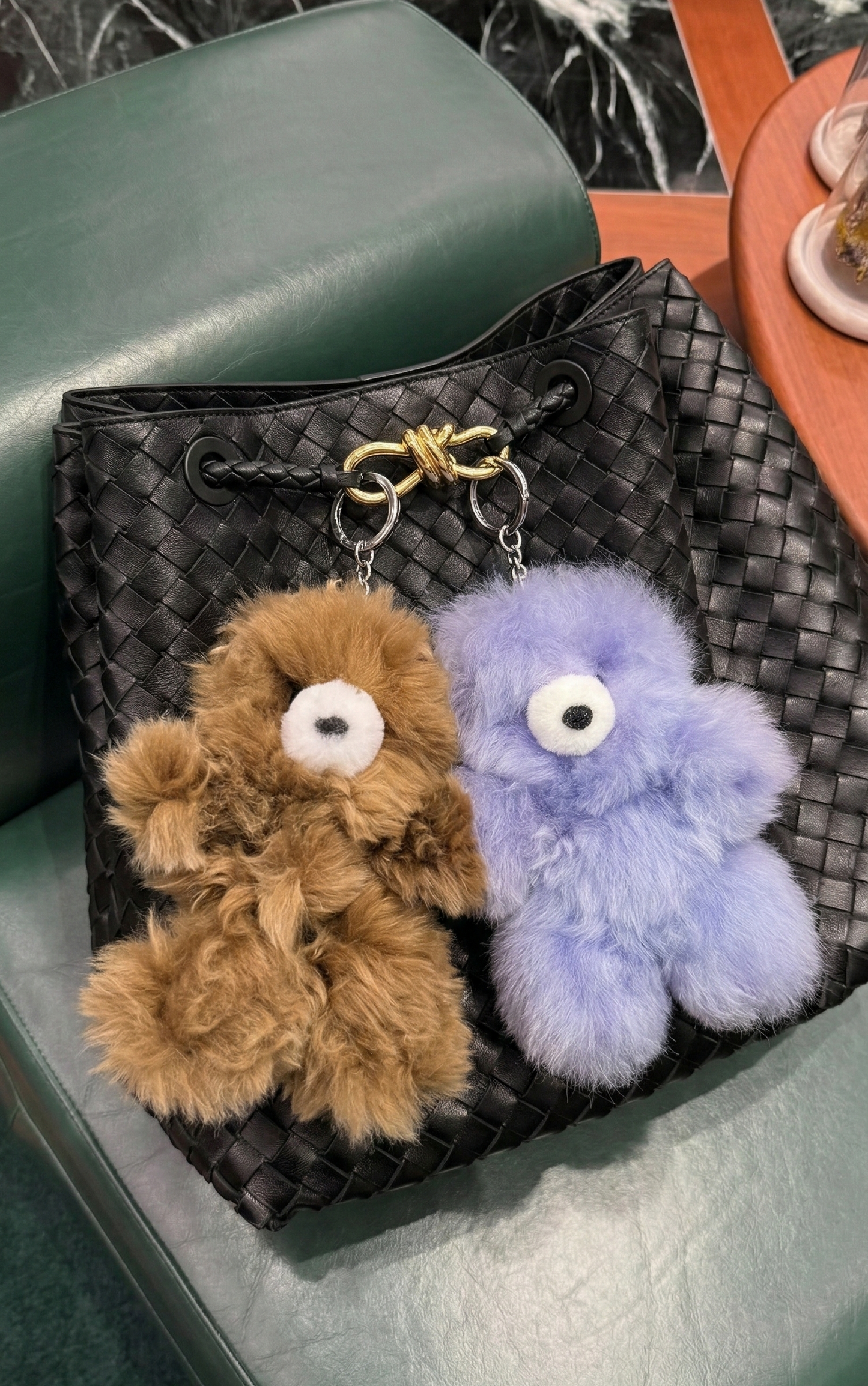 Brown and lavender purple alpaca bear bag charms on a black intrecciato-style woven leather bag on a green velvet chair