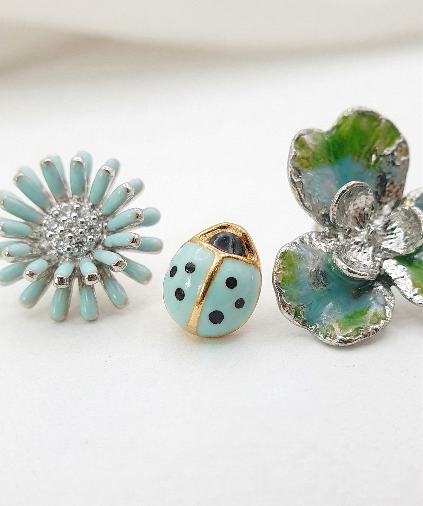 Macro shot of the Fresh Mint 3-piece earring set: a detailed mint daisy, a mint ladybug, and a silver-blue clover on a white surface.