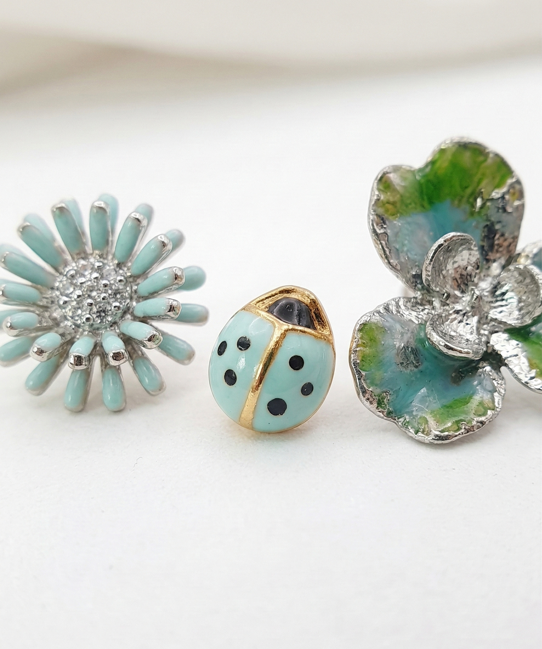 Macro shot of the Fresh Mint 3-piece earring set: a detailed mint daisy, a mint ladybug, and a silver-blue clover on a white surface.