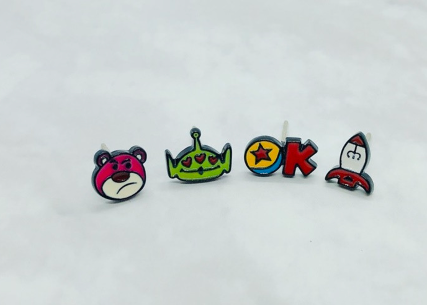 Flat lay of all four Toy Story character earrings - Lotso, Alien, Rocket and Pixar Ball
