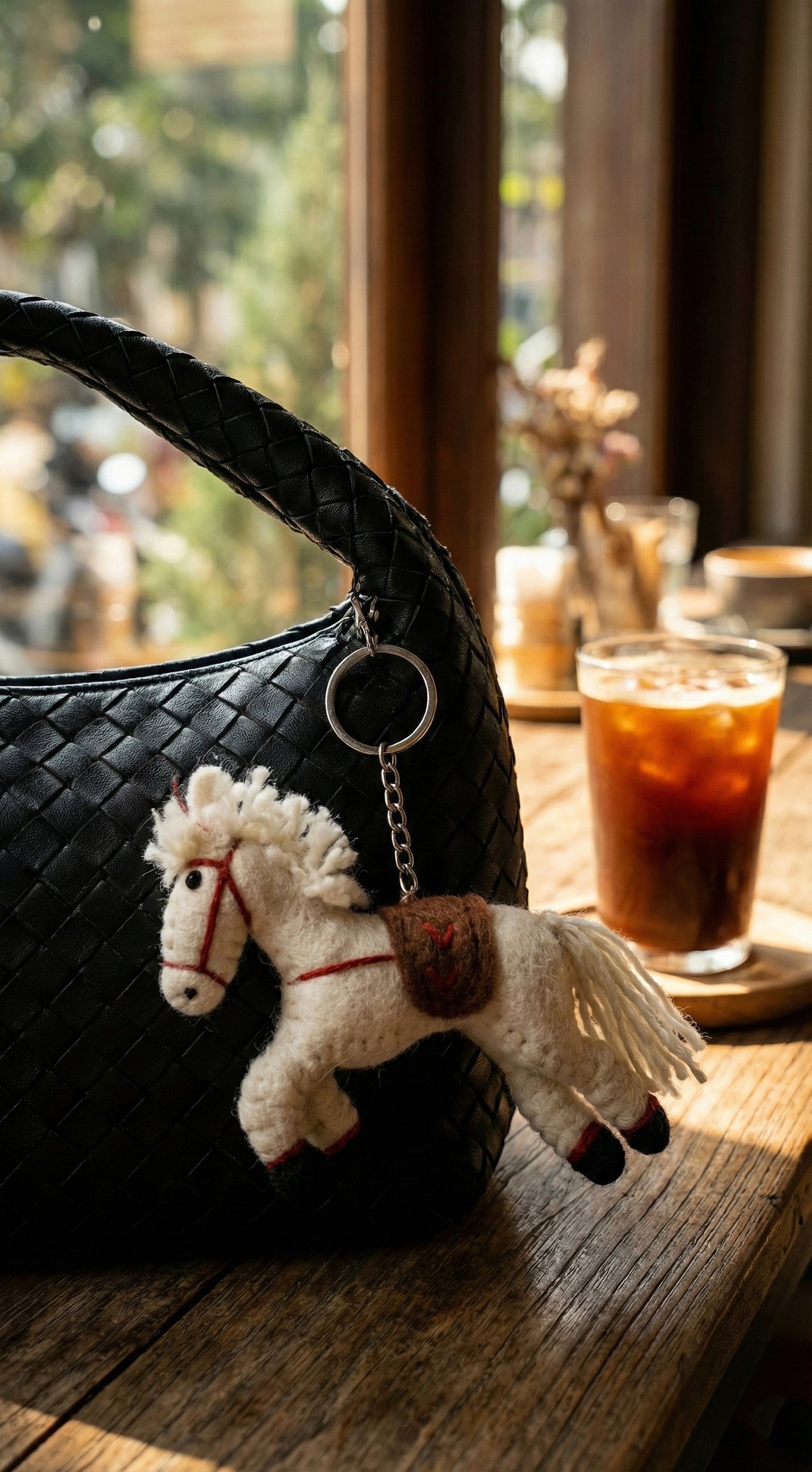 Wool felt horse keychain attached to black woven bag at cafe with iced coffee