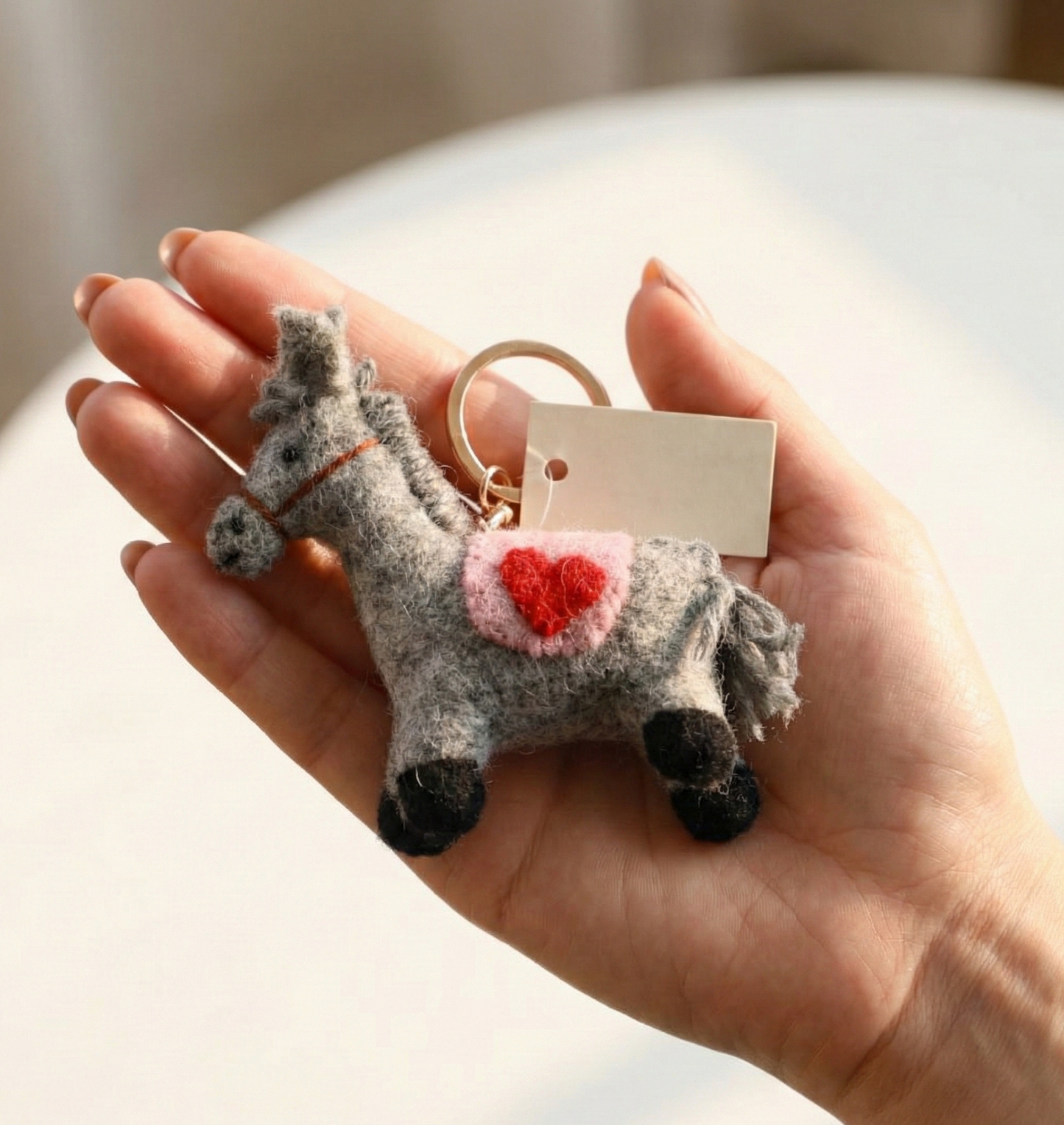 Grey felt horse keychain with pink heart detail held in hand with gold ring