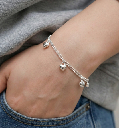 Close-up of a dainty 925 silver heart charm bracelet worn on a wrist against beige linen top and blue denim jeans, casual everyday layered style