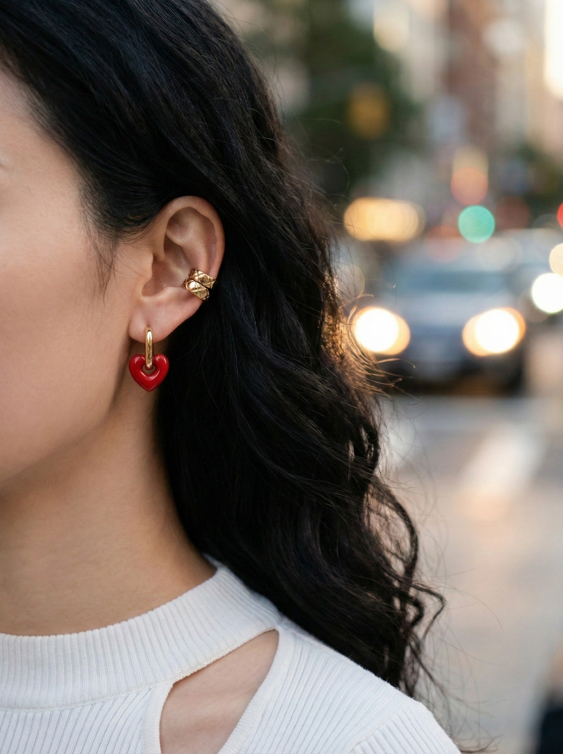 Side profile of woman wearing red heart drop earrings with white knit top, urban street background