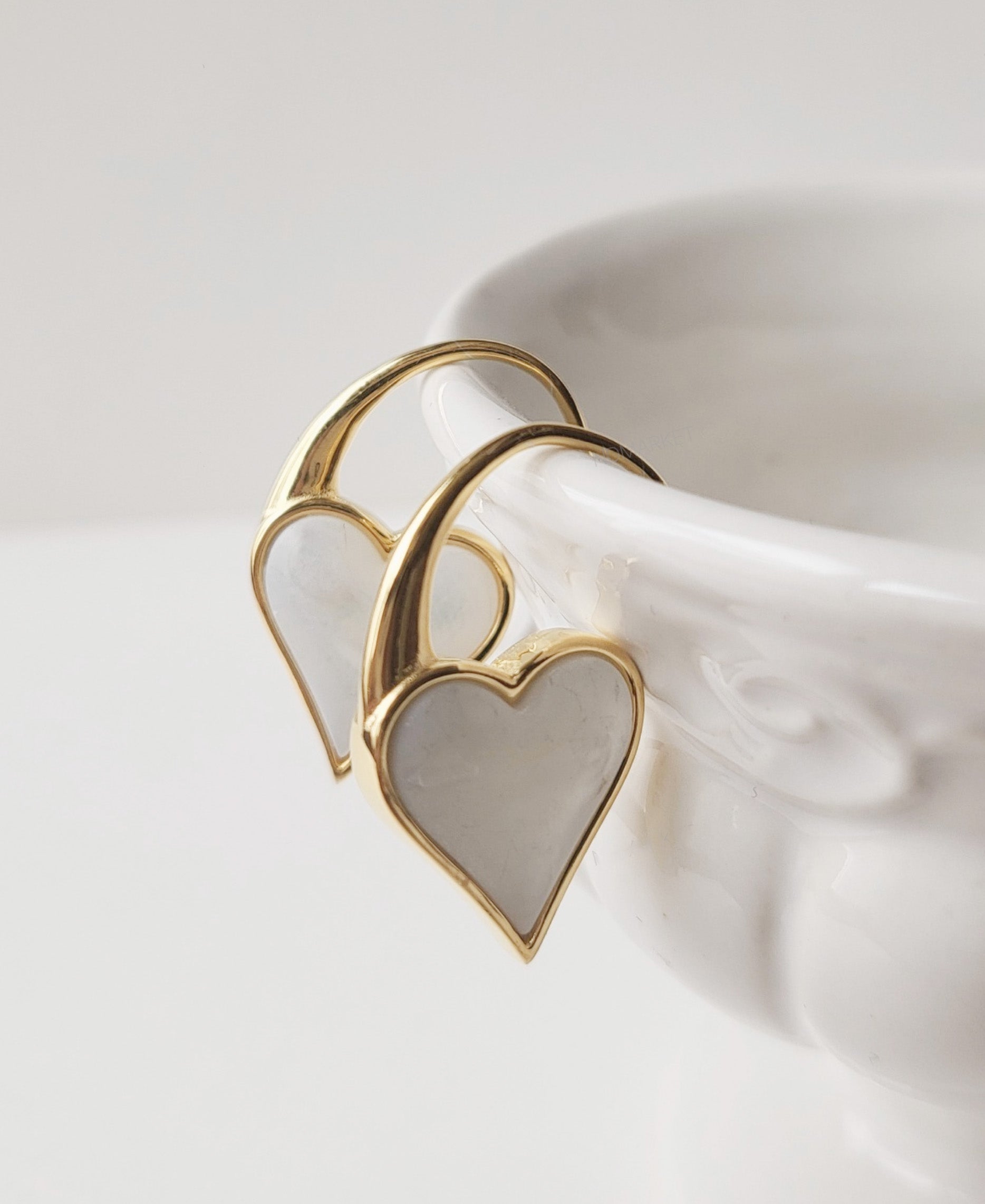 A pair of gold heart hook earrings with mother-of-pearl pendants hanging gracefully on the edge of a white ceramic bowl.
