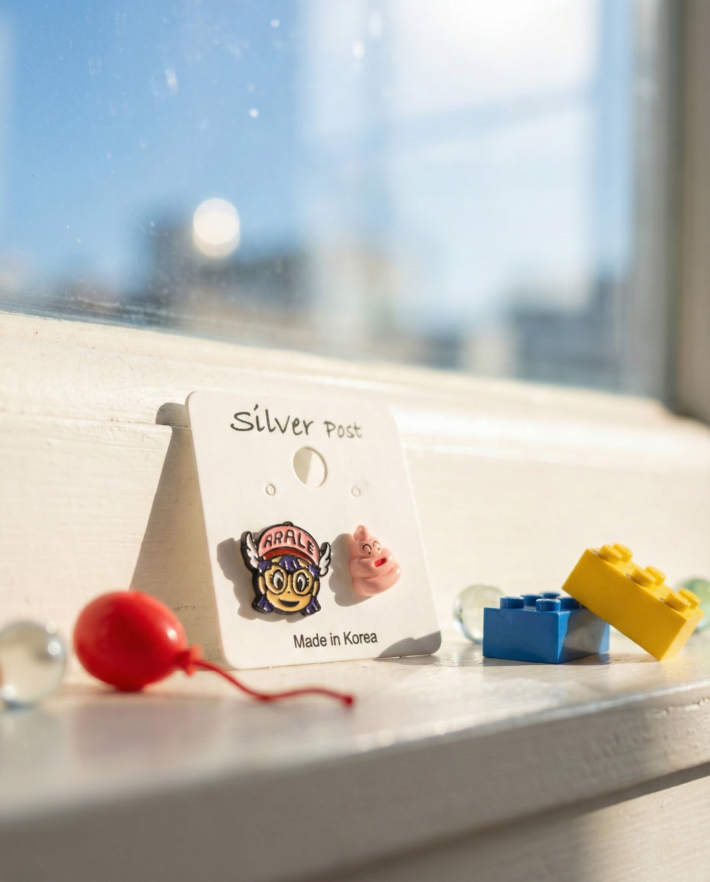 Mismatched anime earrings featuring Arale character with winged cap and cute pink poop, hypoallergenic silver post studs on white card