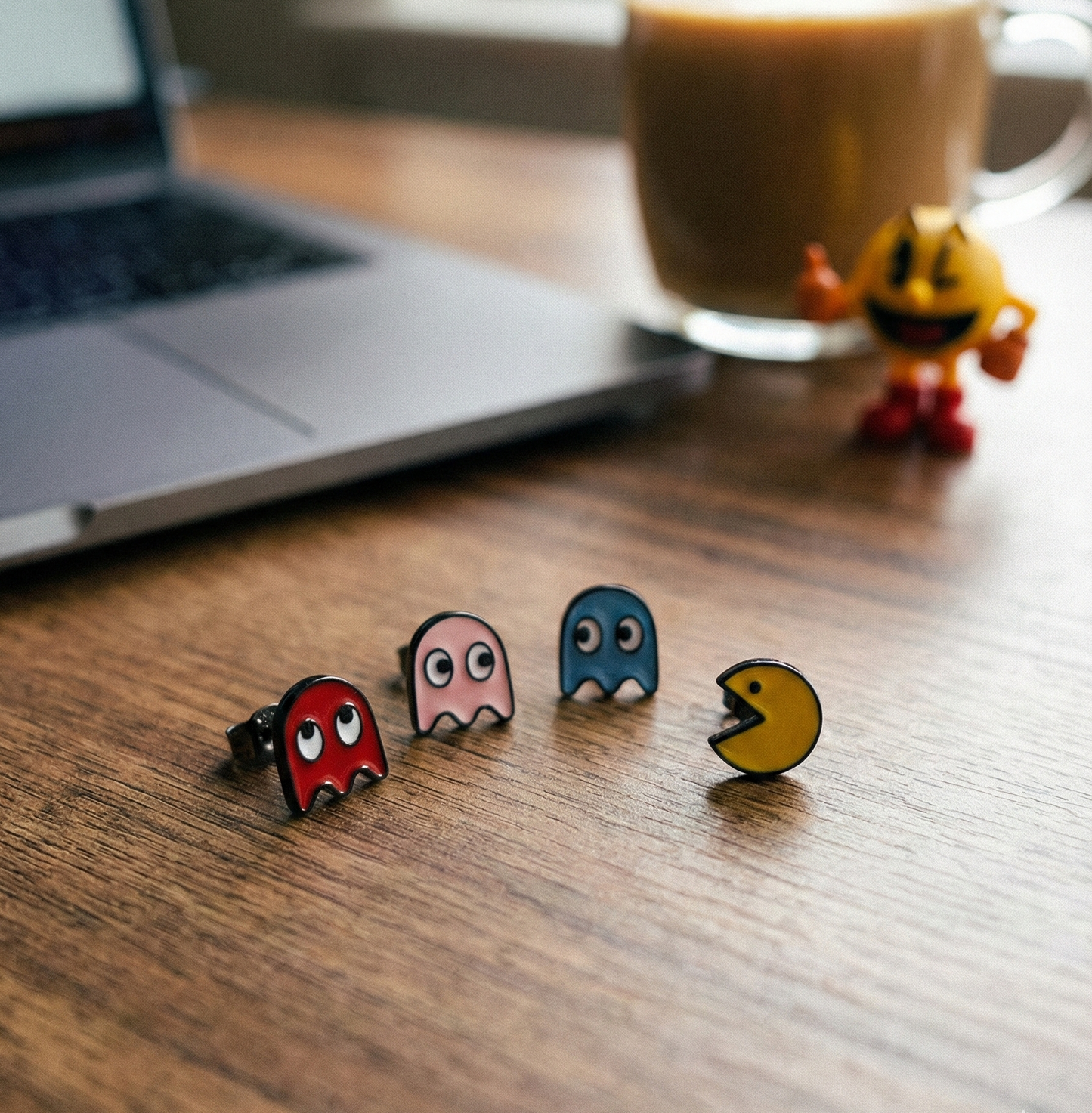 Four individual enamel stud earrings laid out on wooden surface — red ghost, pink ghost, blue ghost, and Pac-Man — with laptop and Pac-Man figurine in background