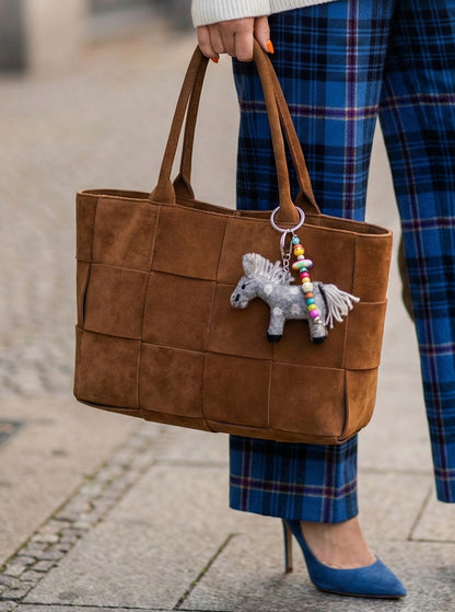 Grey felt donkey keychain with colorful beads hanging on brown suede tote bag held by woman in plaid skirt