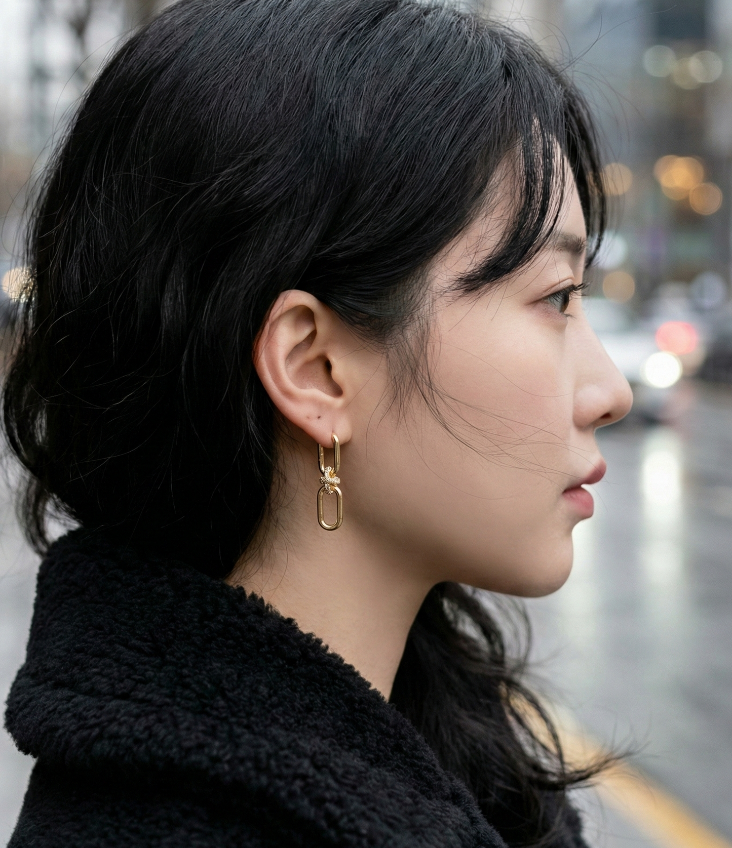 Woman modeling gold pavé knot earrings with black turtleneck sweater, elegant urban look