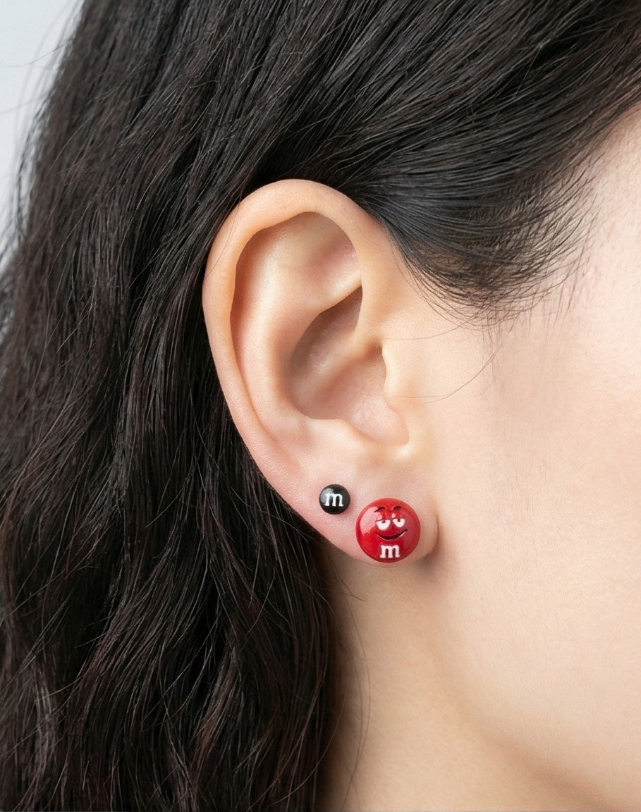 Close-up of M&M's earrings worn in double lobe piercing, red chocolate candy character stud with black mini companion piece, playful everyday earrings, sterling silver hypoallergenic posts, Korean handcrafted