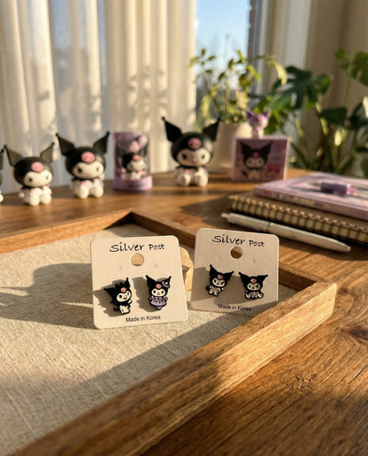 Two pairs of Kuromi earrings on earring cards, placed on a wooden tray in a cozy sunlit room with Kuromi figurines and books in the background.