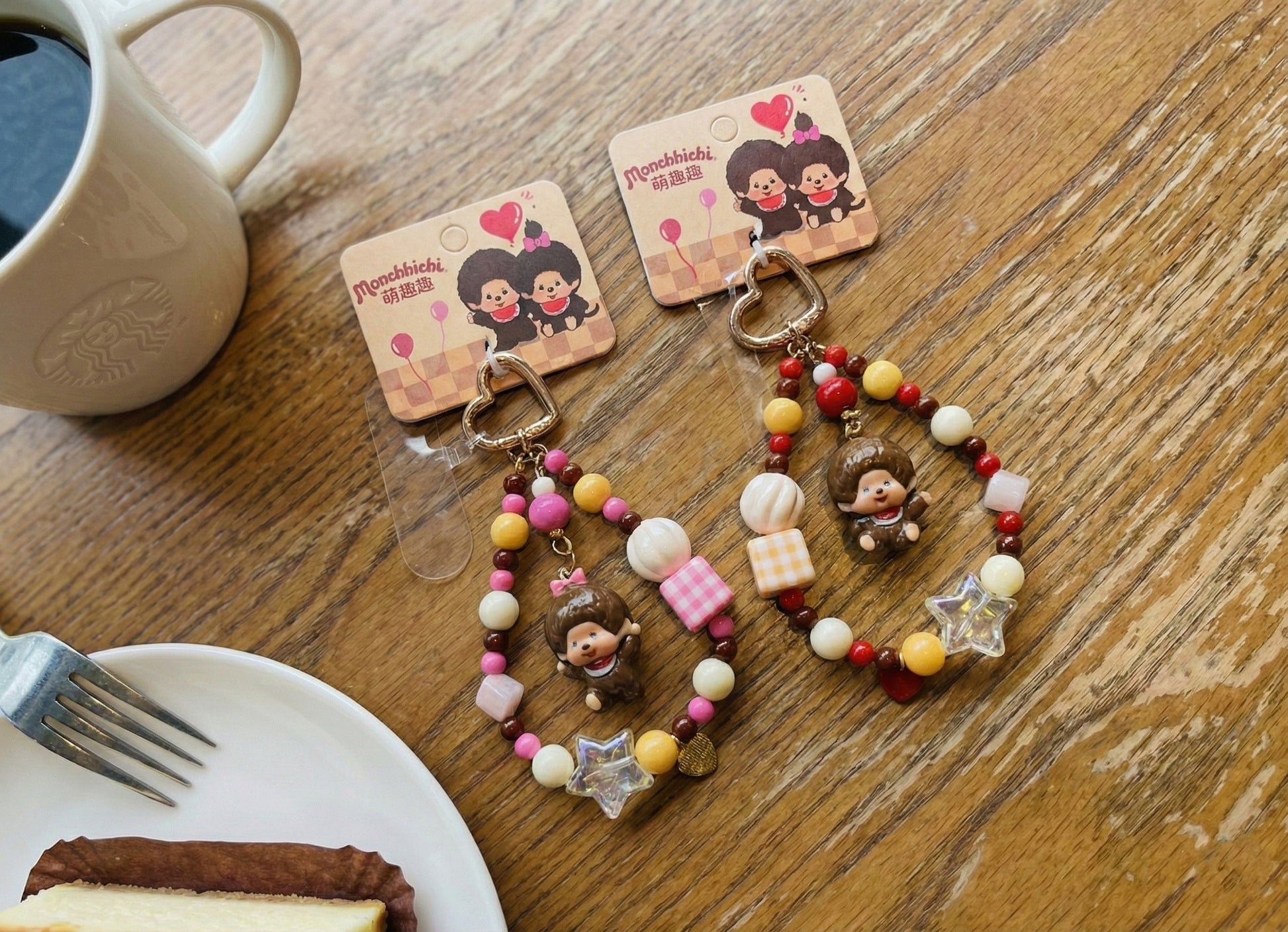 Two handmade Monchhichi bag charms with pink and yellow gingham beads, star accents and heart-shaped gold clasps on wooden cafe table with coffee and dessert