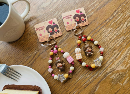 Two handmade Monchhichi bag charms with pink and yellow gingham beads, star accents and heart-shaped gold clasps on wooden cafe table with coffee and dessert