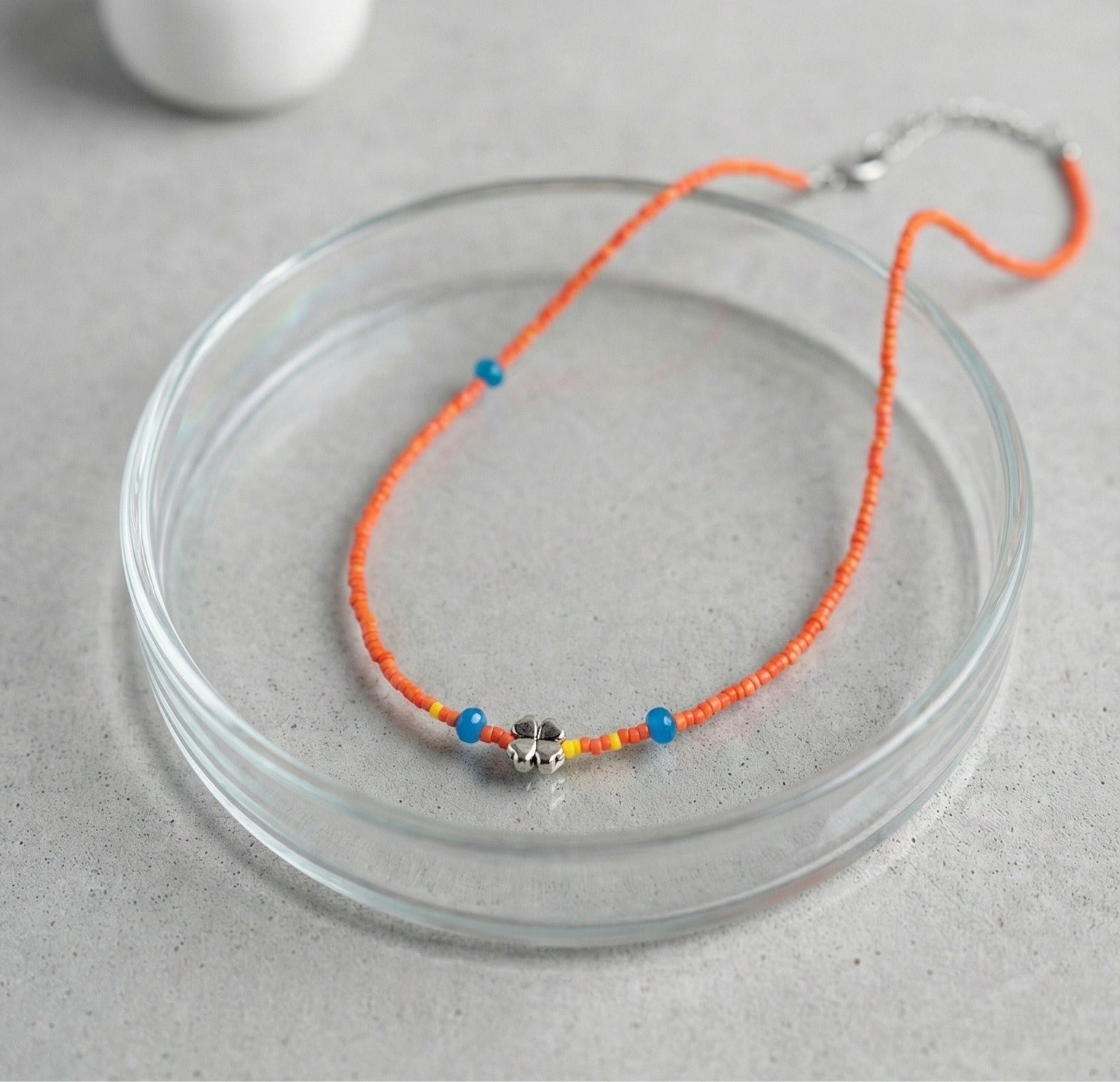 Detail of orange seed bead choker with silver four-leaf clover pendant and blue accent beads in glass bowl