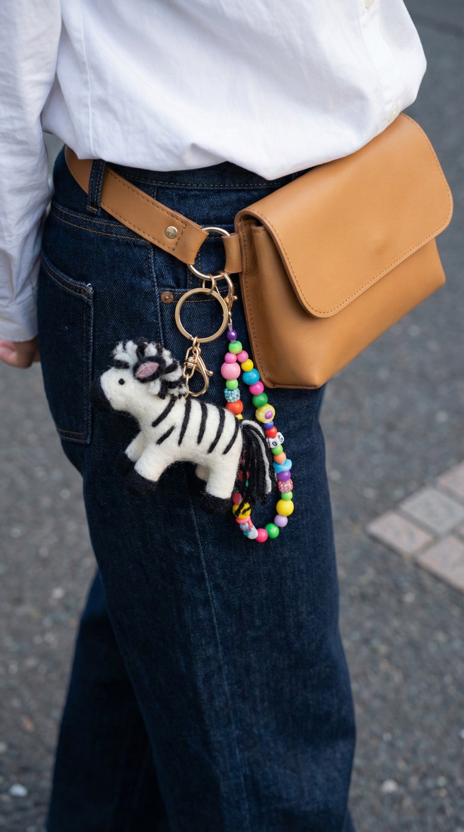 Zebra felt keychain with beads attached to tan crossbody bag worn with white shirt and denim skirt