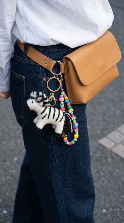 Zebra felt keychain with beads attached to tan crossbody bag worn with white shirt and denim skirt
