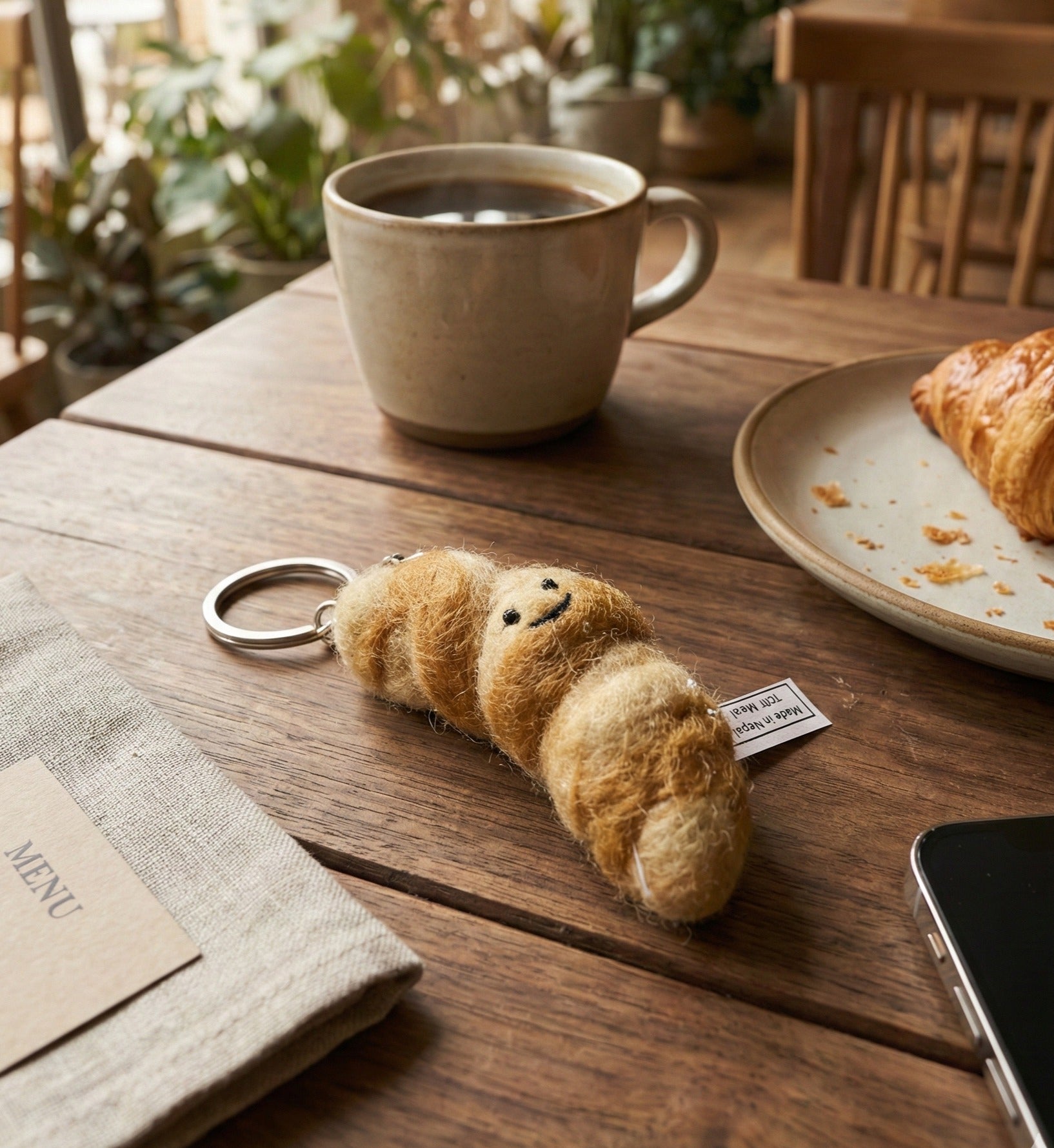 Golden brown felt croissant keychain on wooden cafe table next to coffee and real croissant
